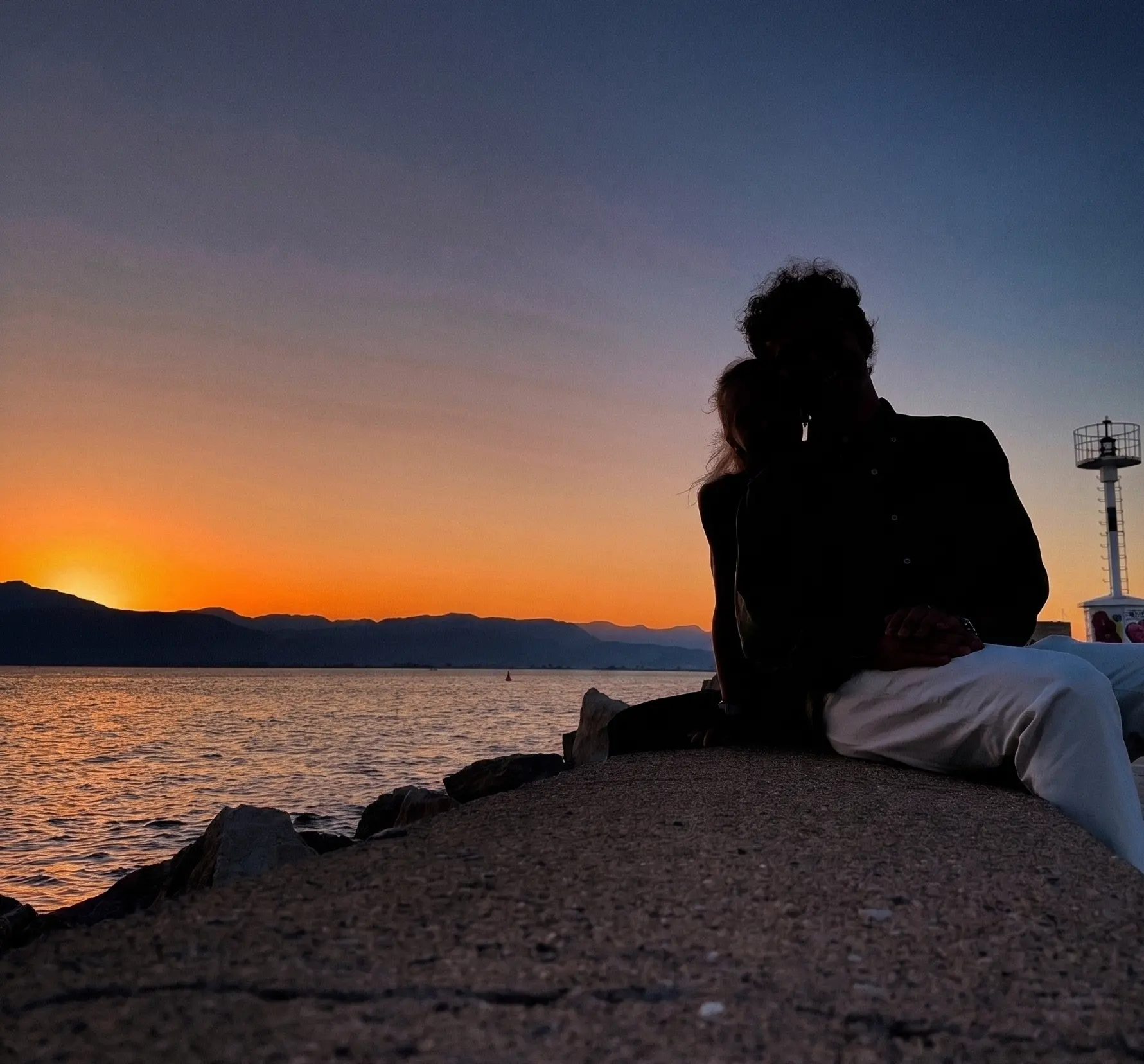 Noi due di spalle, abbracciati al tramonto sul lungomare: silhouette contro il cielo arancione, davanti al mare e al faro.