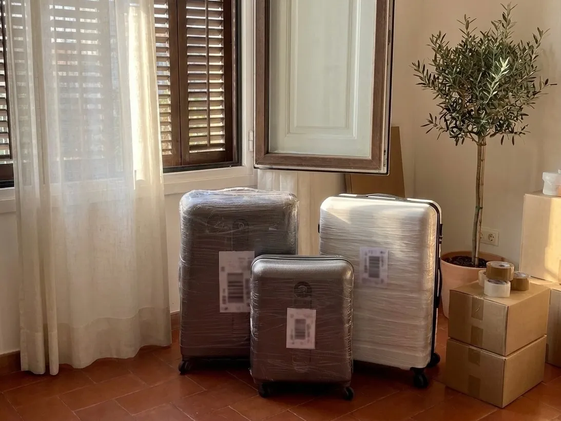 Tre valigie rigide fasciate nel cellophane, ancora con i cartellini del negozio, accanto a scatoloni chiusi col nastro e a un ulivo in vaso, nella luce della finestra di casa.